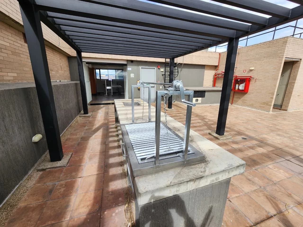 Zona de BBQ en la azotea, con asador de parrilla regulable bajo una pérgola metálica.